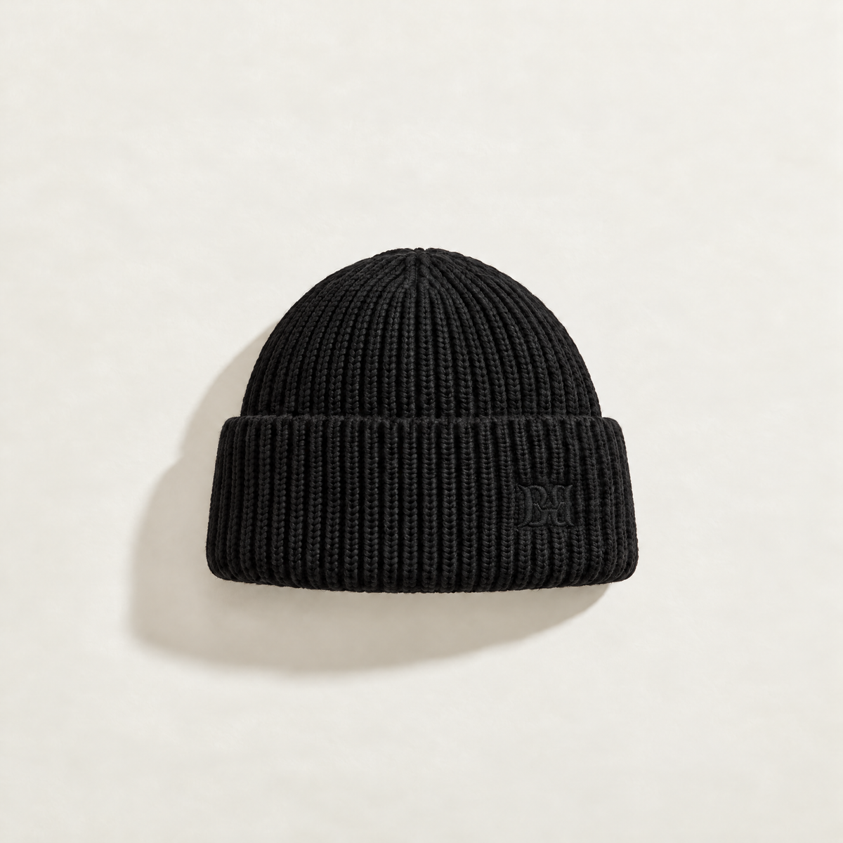 A black ribbed knit beanie hat with a folded cuff, featuring a subtle tonal embroidered logo on the side, displayed on a light beige background.