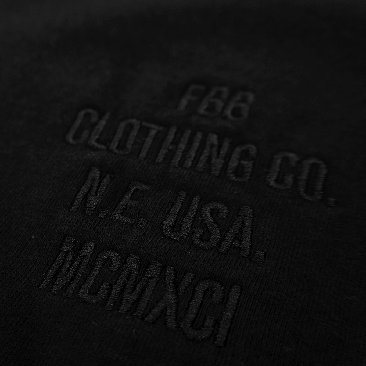 Close-up of a black fabric surface with tonal black embroidery reading 'FBB CLOTHING CO. N.E. USA. MCMXCI'.