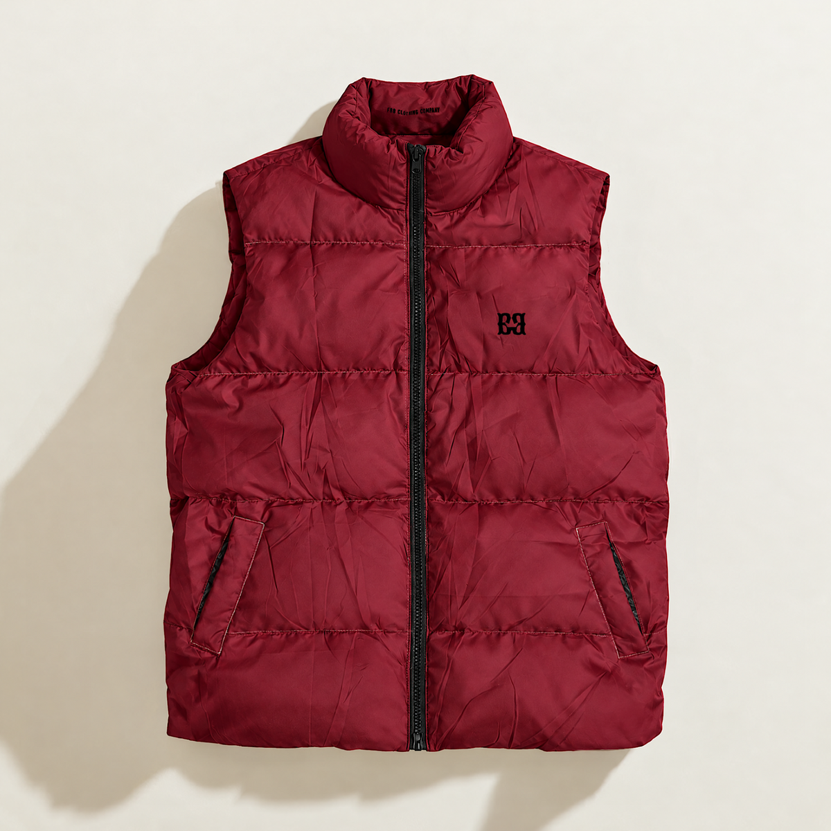 A sleeveless quilted down puffer vest in deep burgundy red, featuring a high stand-up collar, a front zipper closure, and a black embroidered monogram logo on the left chest, displayed flat against a light beige background.