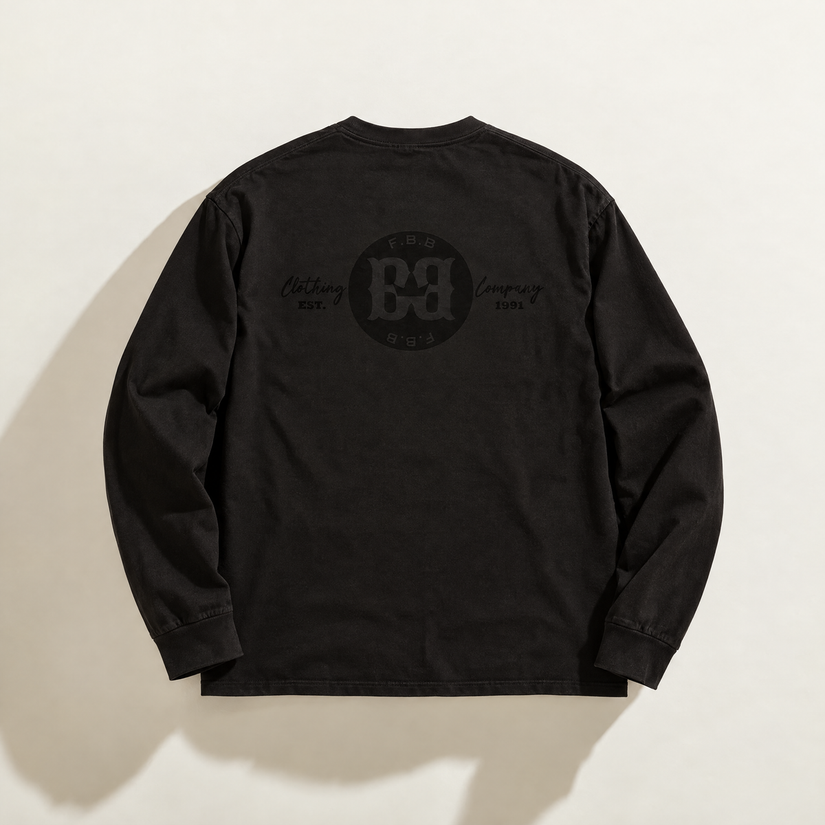 A black long-sleeve t-shirt shown from the back, featuring a tonal black circular print graphic logo from FBB Clothing Company ' and 'Est. 1991' text on the upper back, laid flat against a light beige background.