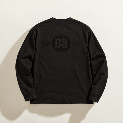 A black long-sleeve t-shirt shown from the back, featuring a tonal black circular print graphic logo from FBB Clothing Company ' and 'Est. 1991' text on the upper back, laid flat against a light beige background.