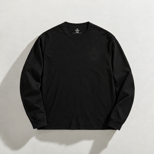 A black long-sleeve heavyweight cotton crewneck t-shirt with a tonal embroidered chest graphic, left cuff and right cuff embroidered ensignia laid flat on a light beige background.