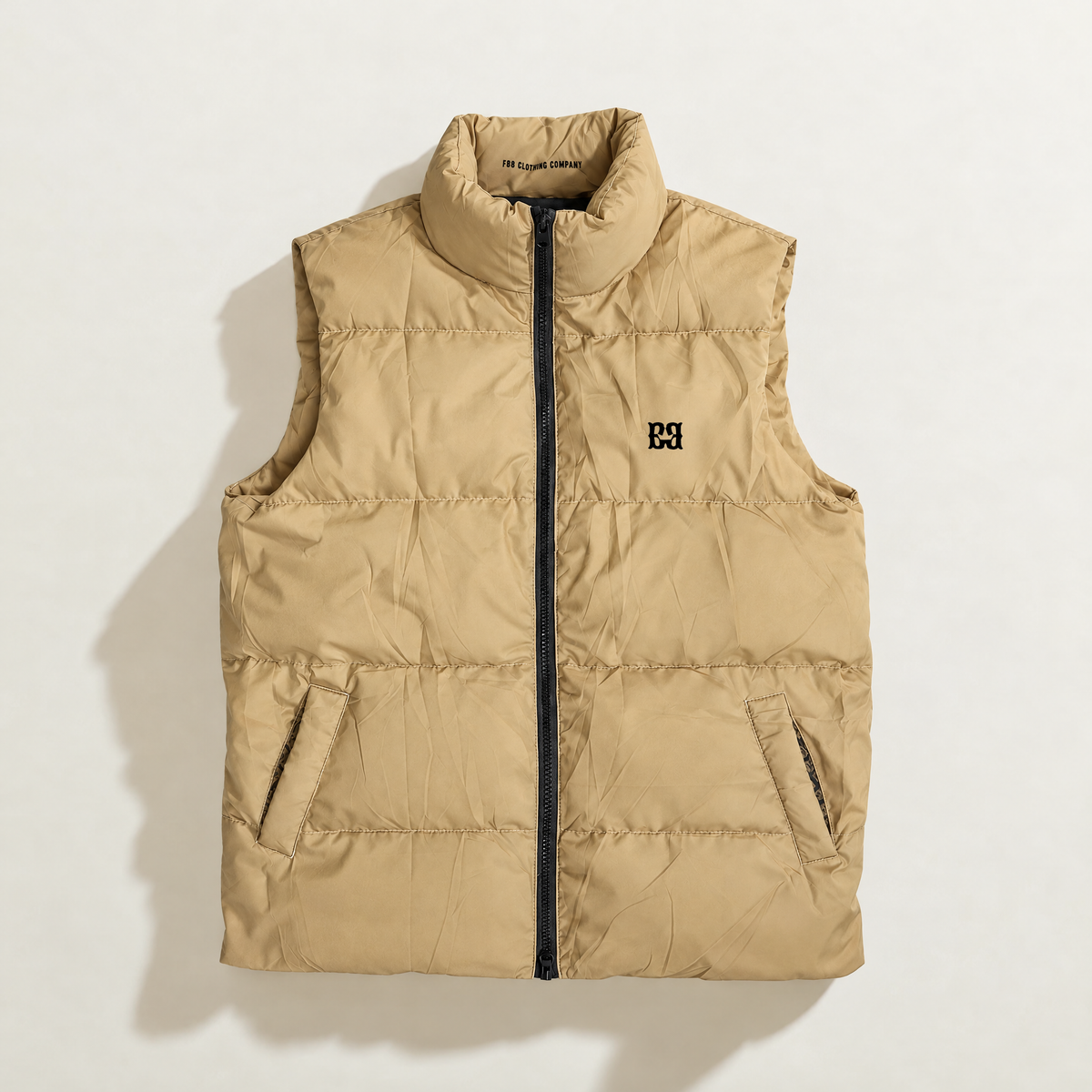 A beige quilted down puffer vest with a black front zipper, black logo on the left chest, and zippered hand pockets, laid flat against a white background.