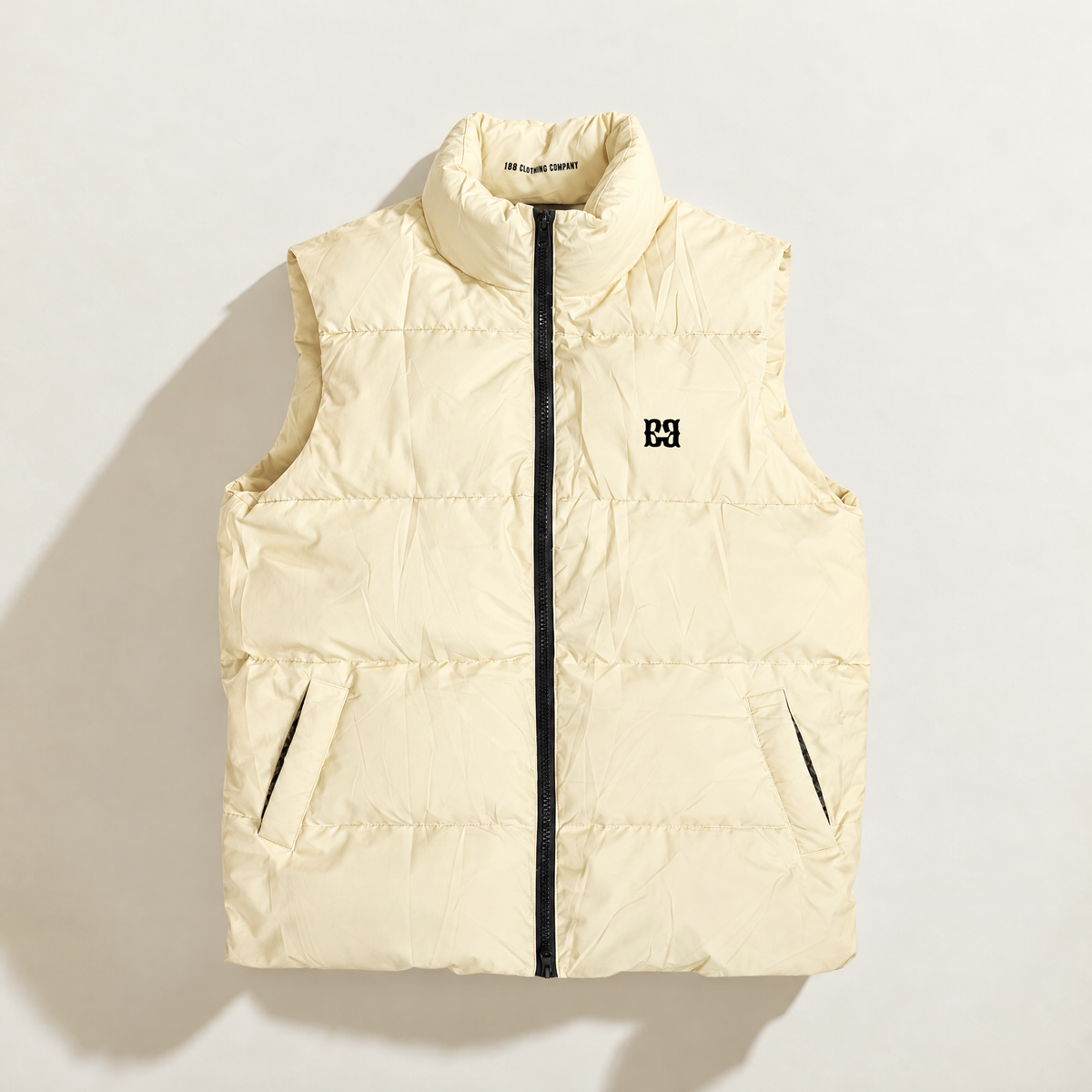 A sleeveless quilted down puffer vest in vanilla cream, featuring a black front zipper, a stand-up collar, and a small black logo on the left chest, displayed against a solid white background.