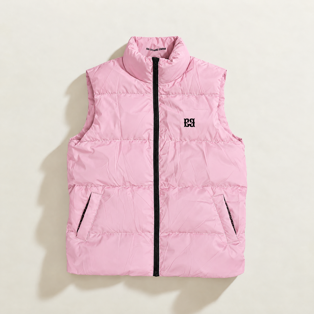 A light pink lavender sleeveless down puffer vest with a high stand-up collar, black front zipper, and a small black embroidered logo on the left chest, laid flat on a light beige background.